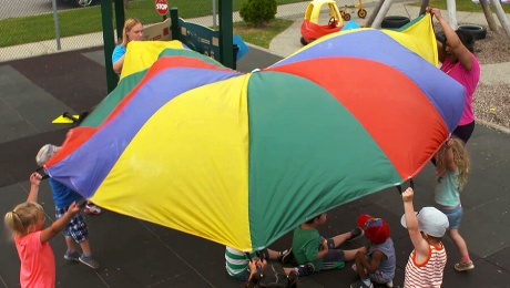 Children and caregivers playing with a multicoloured parachute outside at a day care facility.