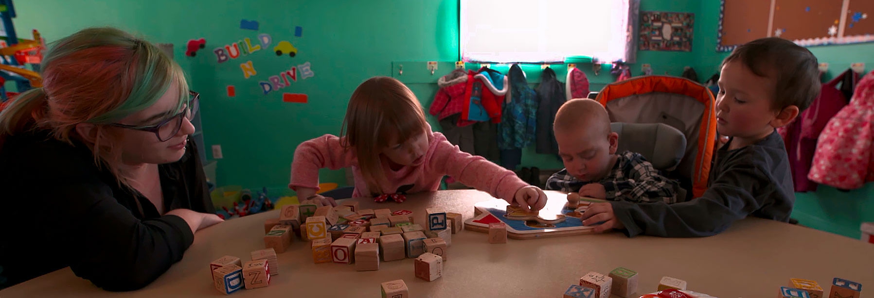 Children playing at a table with blocks while a care giver gives encouragement.