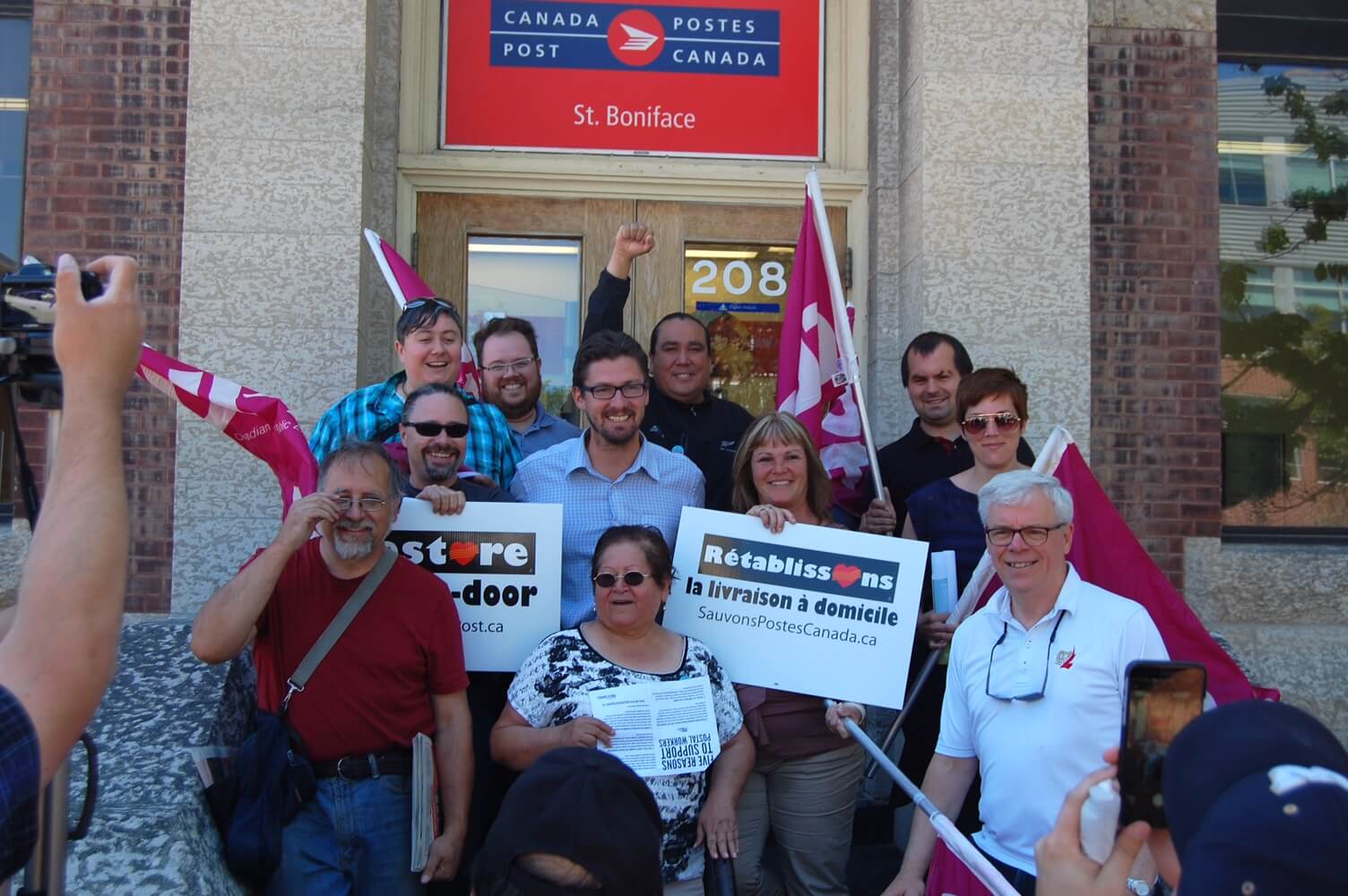 Groupe de personnes devant le bureau de poste de Saint-Boniface, certaines tenant une affiche « Rétablissons la livraison à domicile ».  